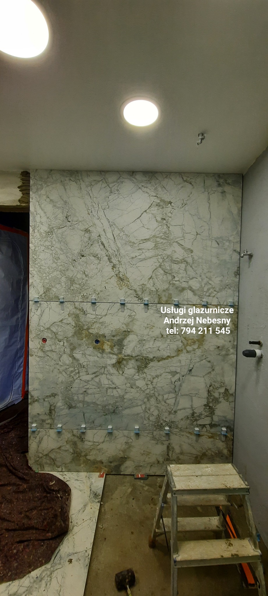 Układanie wielkoformatowych płytek imitujących marmur na ścianie, widoczne krzyżyki dystansowe, fragment drabiny i gumowy młotek.