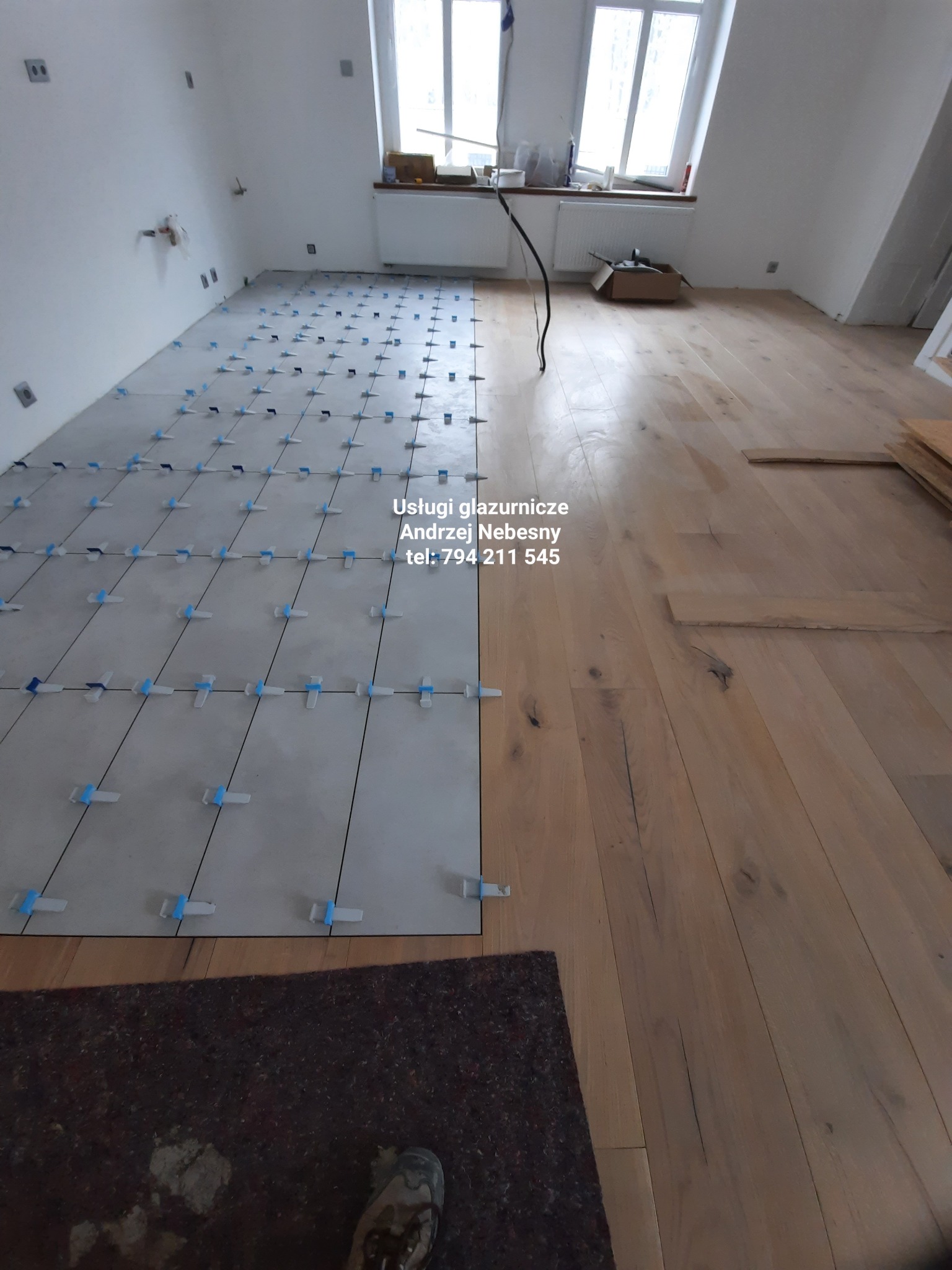 Połowa podłogi wyłożona jasnymi płytkami z niebieskimi krzyżykami dystansowymi, druga połowa z drewnianą podłogą w trakcie remontu, widoczne okno i fragment buta na dywanie.