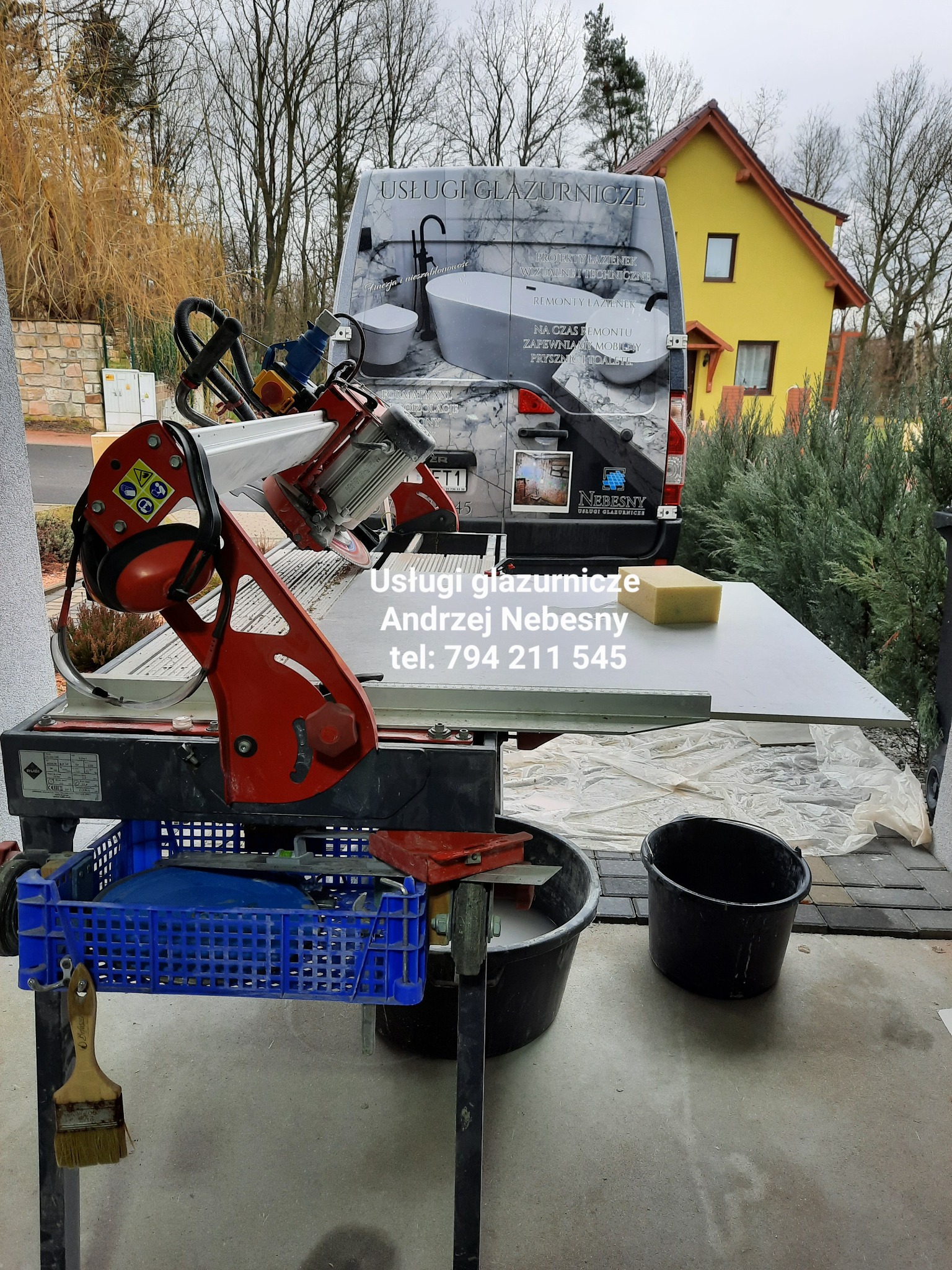 Elektryczna przecinarka do płytek z tarczą diamentową, przygotowana do cięcia dużej płyty, obok wiaderko z wodą, w tle samochód z reklamą usług glazurniczych i żółty dom