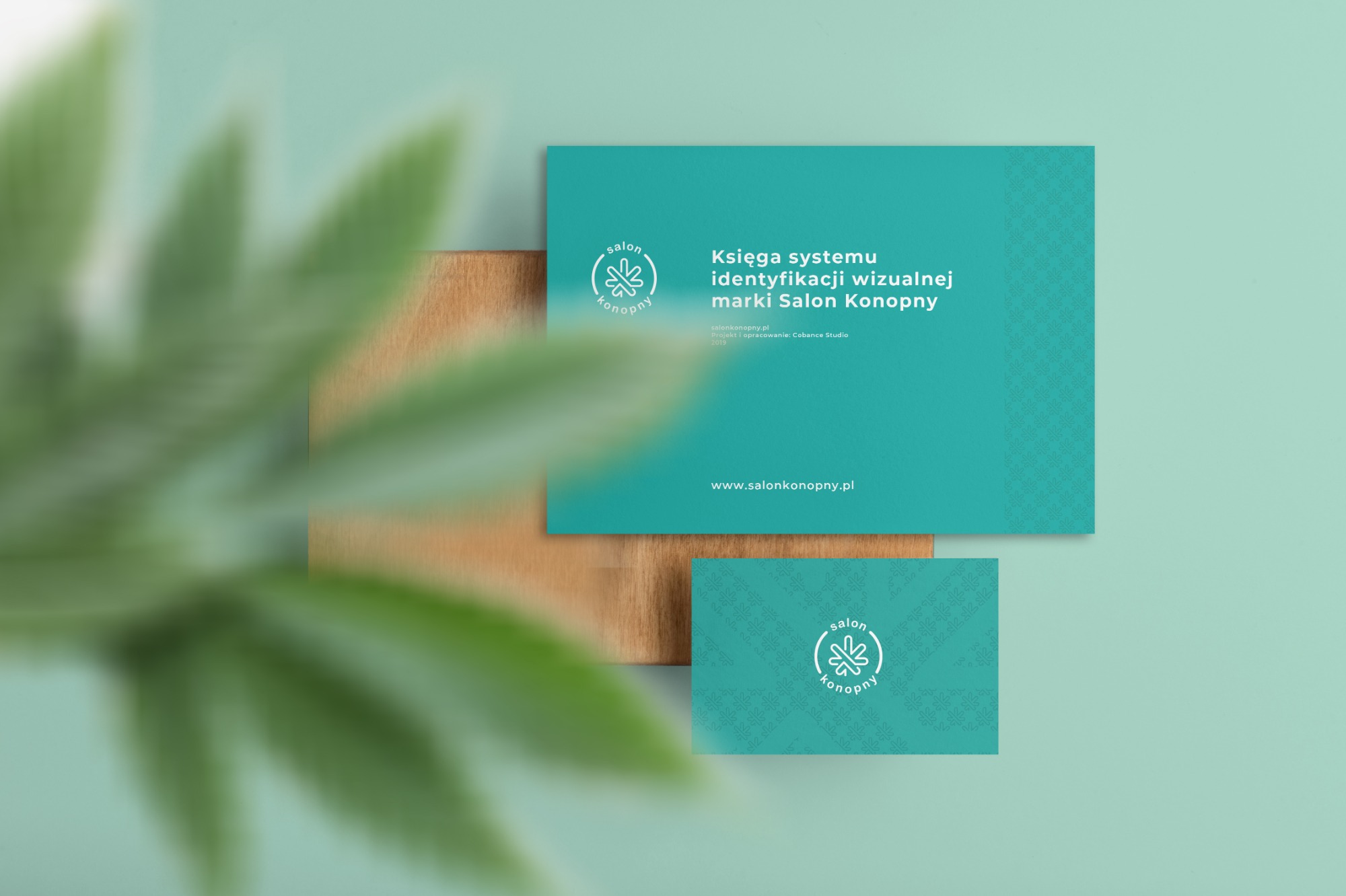 Księga identyfikacji wizualnej marki Salon Konopny w kolorze turkusowym, umieszczona na drewnianej podstawce. W tle rozmyte liście rośliny.