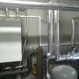 Wnętrze kotłowni z kotłem gazowym, rurami w izolacji termicznej i zbiornikiem wyrównawczym, widoczne manometry i zawory.