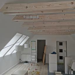 Usługi remontowo budowlane - Poddasze w trakcie prac wykończeniowych: widoczne drewniane belki sufitowe z zamontowanym oświetleniem, białe ściany, okno dachowe wpuszczające światło, drabina, białe meble w kartonach i folia...