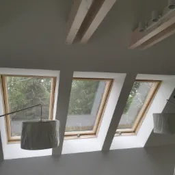 Trzy okna dachowe z drewnianą ramą w białym pokoju na poddaszu, widok na zielone drzewa za oknem, drewniane belki na suficie i nowoczesne oświetlenie.