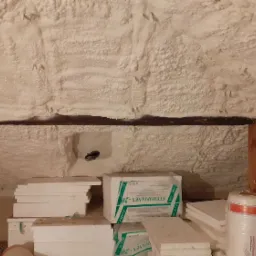 Poddasze po natrysku pianki izolacyjnej, widoczne elementy drewnianej konstrukcji oraz stos płyt styropianowych i rolki materiału izolacyjnego w folii.