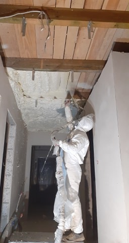 Pracownik w kombinezonie ochronnym aplikuje izolację natryskową na sufit poddasza, widoczne drewniane belki stropowe i przewody elektryczne.