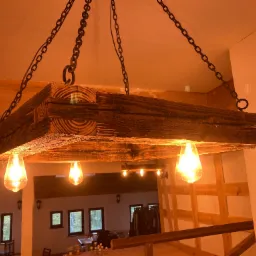 Rustykalna lampa wisząca z grubego, ciemnego drewna, podwieszona na łańcuchach, z żarówkami w stylu retro, oświetlająca wnętrze z drewnianymi elementami konstrukcyjnymi.