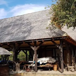 Drewniany budynek z gontowym dachem, wsparty na drewnianych kolumnach, z widocznymi narzędziami i materiałami w środku, otoczony zielenią.