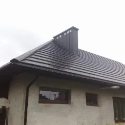 Dom jednorodzinny z ciemną dachówką ceramiczną, czarną rynną i kominem, widoczne okna z brązowymi ramami i fragment boiska do koszykówki.