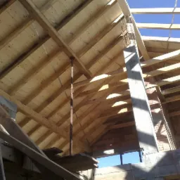 Wnętrze dachu w trakcie budowy, widoczna drewniana więźba dachowa z deskowaniem, podpora teleskopowa i betonowy filar na tle błękitnego nieba.