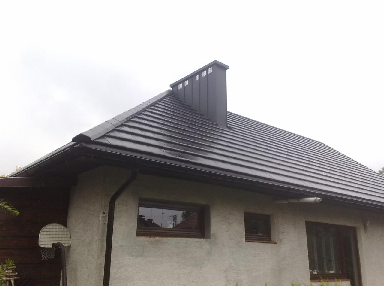 Dom jednorodzinny z ciemną dachówką ceramiczną, czarną rynną i kominem, widoczne okna z brązowymi ramami i fragment boiska do koszykówki.