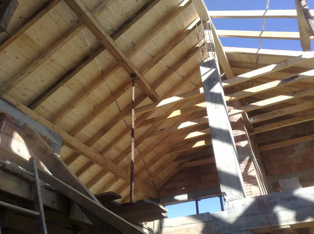 Wnętrze dachu w trakcie budowy, widoczna drewniana więźba dachowa z deskowaniem, podpora teleskopowa i betonowy filar na tle błękitnego nieba.
