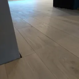 Ułożone panele podłogowe w jasnym odcieniu drewna, widoczne wzdłuż ściany z widoczną szczeliną dylatacyjną, z fragmentem czarnej ściany w tle.