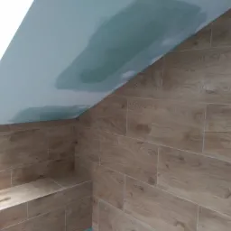 Poddasze z wykończoną ścianą i siedziskiem z płytek imitujących drewno, sufit częściowo pokryty gładzią szpachlową.