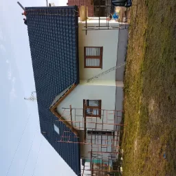 Dom jednorodzinny z częściowo położonym ciemnym dachem, elewacja w jasnym kolorze, widoczne rusztowania i drabina oparta o ścianę, dwóch pracowników na dachu.