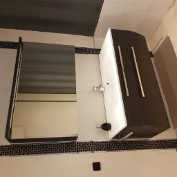 Nowoczesna łazienka z umywalką w szafce z ciemnego drewna, lustrem w szarej ramie i dekoracyjną mozaiką wzdłuż ściany.