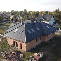 Dariusz Fojcik Firma handlowo usługowa Darplex - Dom w stanie surowym otwartym z ciemną dachówką, widoczne okna dachowe, na pierwszym planie materiały budowlane i betoniarka.