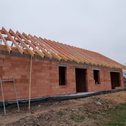 Dariusz Fojcik Firma handlowo usługowa Darplex - Budowa domu murowanego z czerwonej cegły: wzniesione ściany z otworami okiennymi, częściowo wykonana drewniana więźba dachowa, widoczne rusztowania i teren budowy w pochmurny dzień.