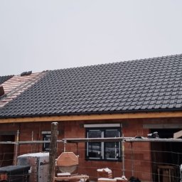 Dariusz Fojcik Firma handlowo usługowa Darplex - Montaż czarnej dachówki na nowym domu z czerwonej cegły, widoczna drabina i częściowo pokryty dach w zimowej scenerii.