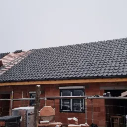 Montaż czarnej dachówki na nowym domu z czerwonej cegły, widoczna drabina i częściowo pokryty dach w zimowej scenerii.