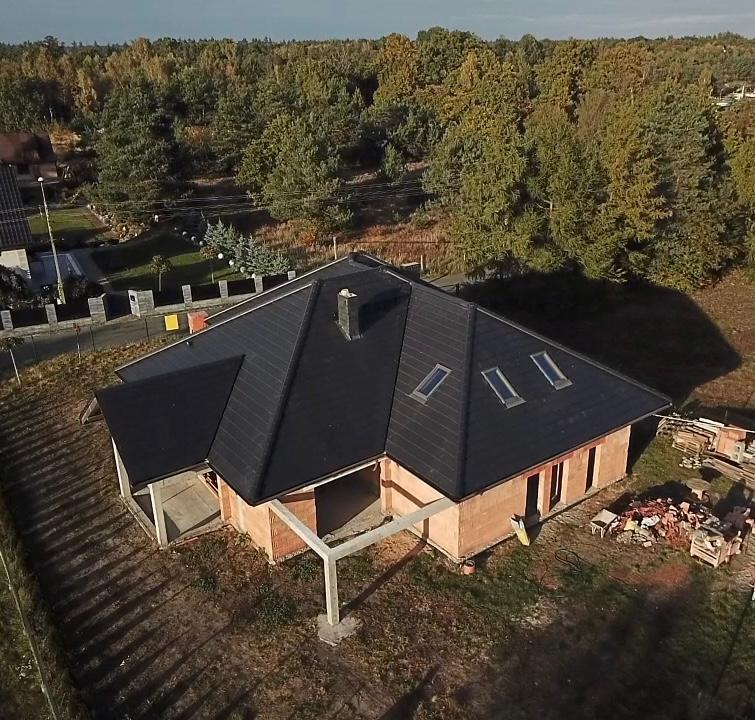 Nowoczesny dom w trakcie budowy z czarnym dachem pokrytym dachówką, widoczne okna dachowe, ściany z cegły, betonowe podpory werandy i stos materiałów budowlanych obok.