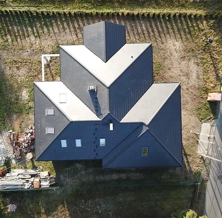 Widok z góry na dom z ciemnym, wielospadowym dachem pokrytym gontem bitumicznym, z widocznymi oknami dachowymi i kominem. Na ziemi leżą materiały budowlane.
