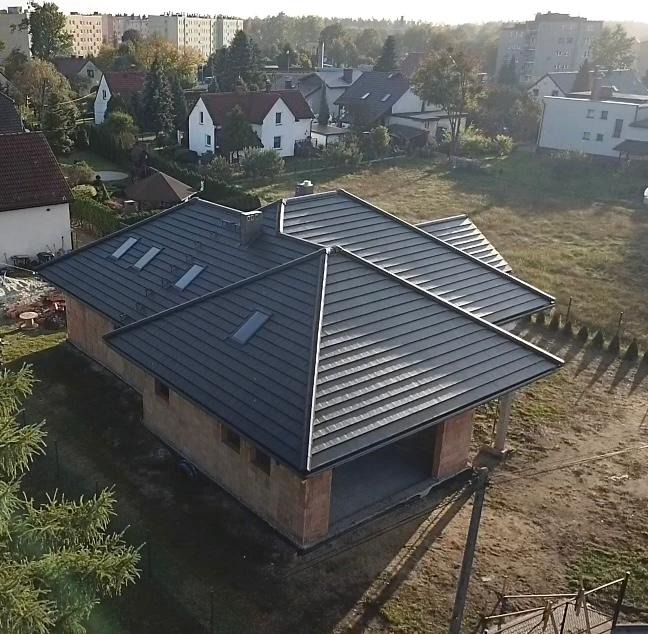 Surowy budynek jednorodzinny z ciemnoszarą, metalową konstrukcją dachową z oknami połaciowymi, w trakcie budowy, z widocznymi elementami konstrukcyjnymi i pustakami ceramicznymi, na tle zabudowy...