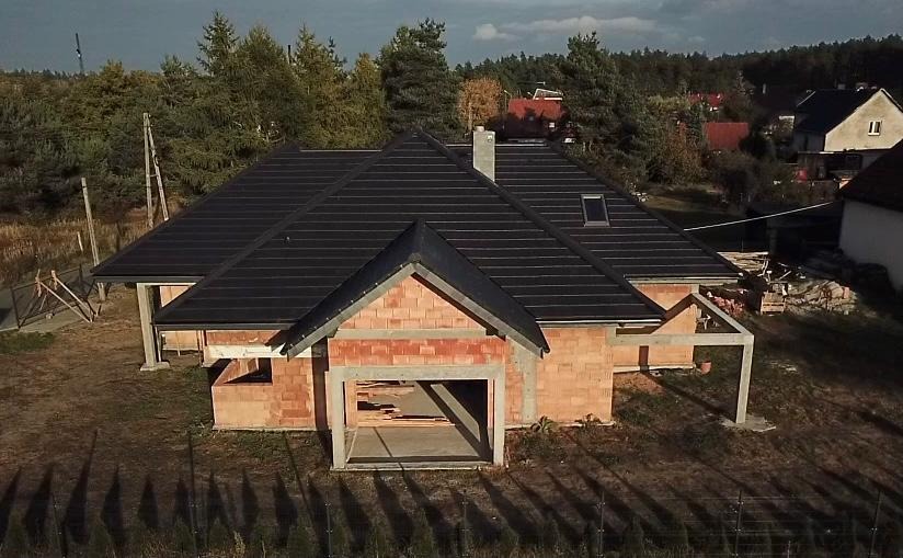 Niedokończony dom z cegły z czarnym dachem w trakcie budowy, widoczne elementy konstrukcyjne garażu i las w tle.