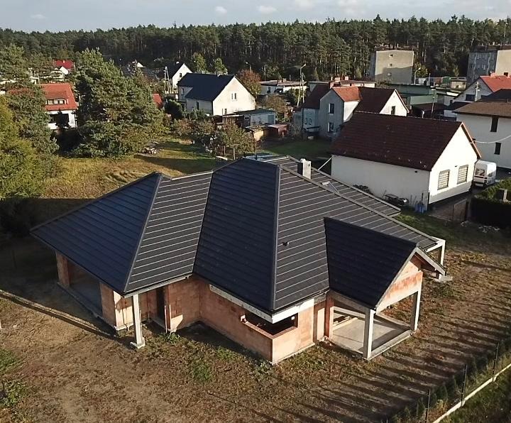 Nowo położony, ciemny dach na surowym domu z czerwonej cegły, widok z góry, w tle zabudowa jednorodzinna i las.