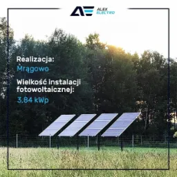 Cztery panele słoneczne w rzędzie na łące, z lasem w tle i logo firmy Alex Electro w rogu. Informacja o realizacji w Mrągowie i mocy instalacji 3.84 kW.