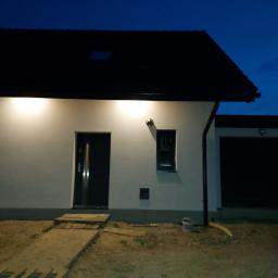 Usługi elektroinstalacyjne i teletechniczne Loranc Piotr - Nowoczesny dom jednorodzinny oświetlony zewnętrznymi lampami ściennymi po zmroku, widoczne okno, drzwi wejściowe i garaż.