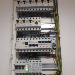 Otwarta skrzynka rozdzielcza z bezpiecznikami i przewodami marki Eaton, widoczne diody LED na jednym z elementów, całość zamontowana na białej ścianie.