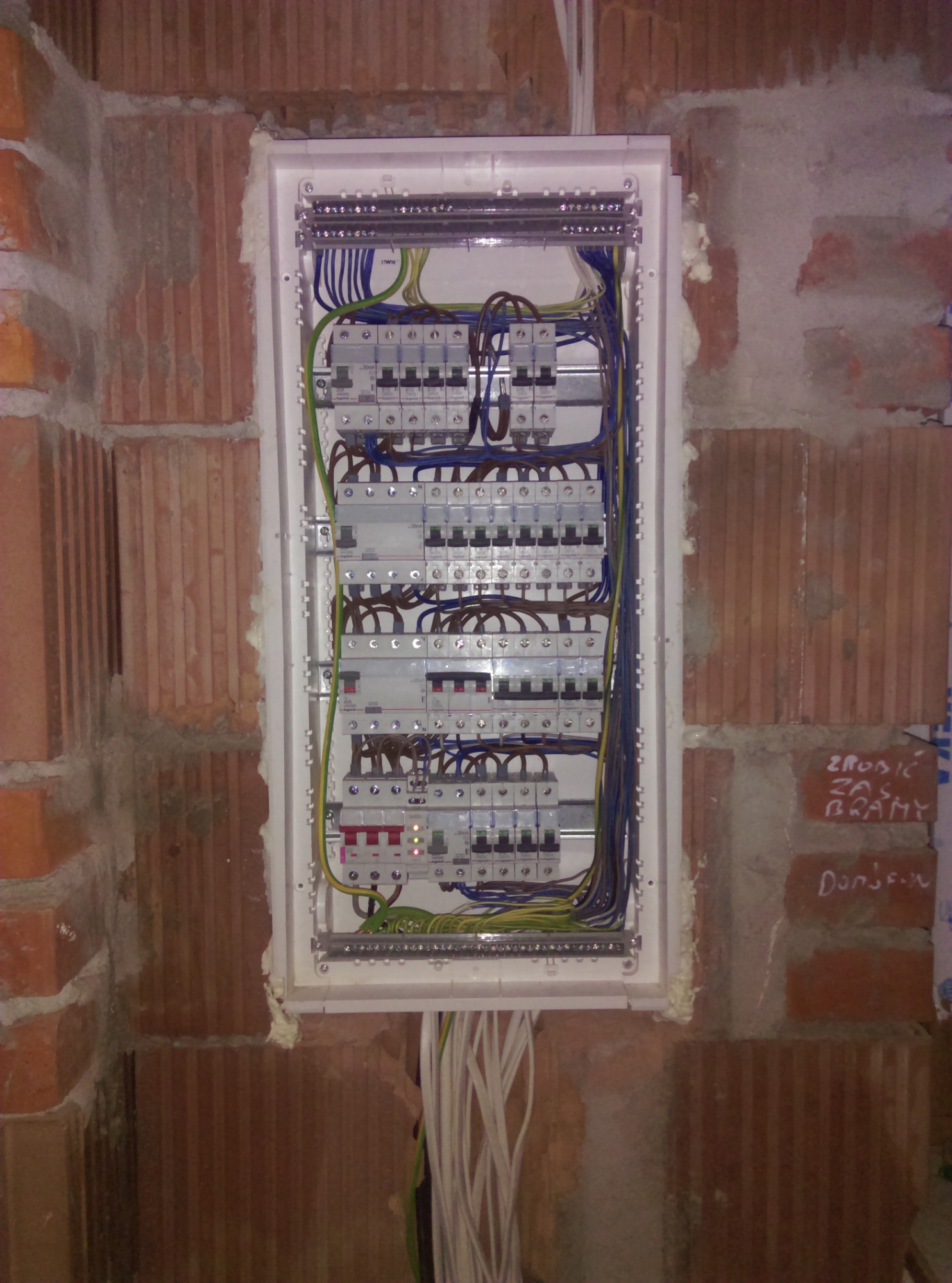 Rozdzielnica elektryczna zamontowana w surowej ścianie z cegły, widoczne zabezpieczenia i okablowanie.