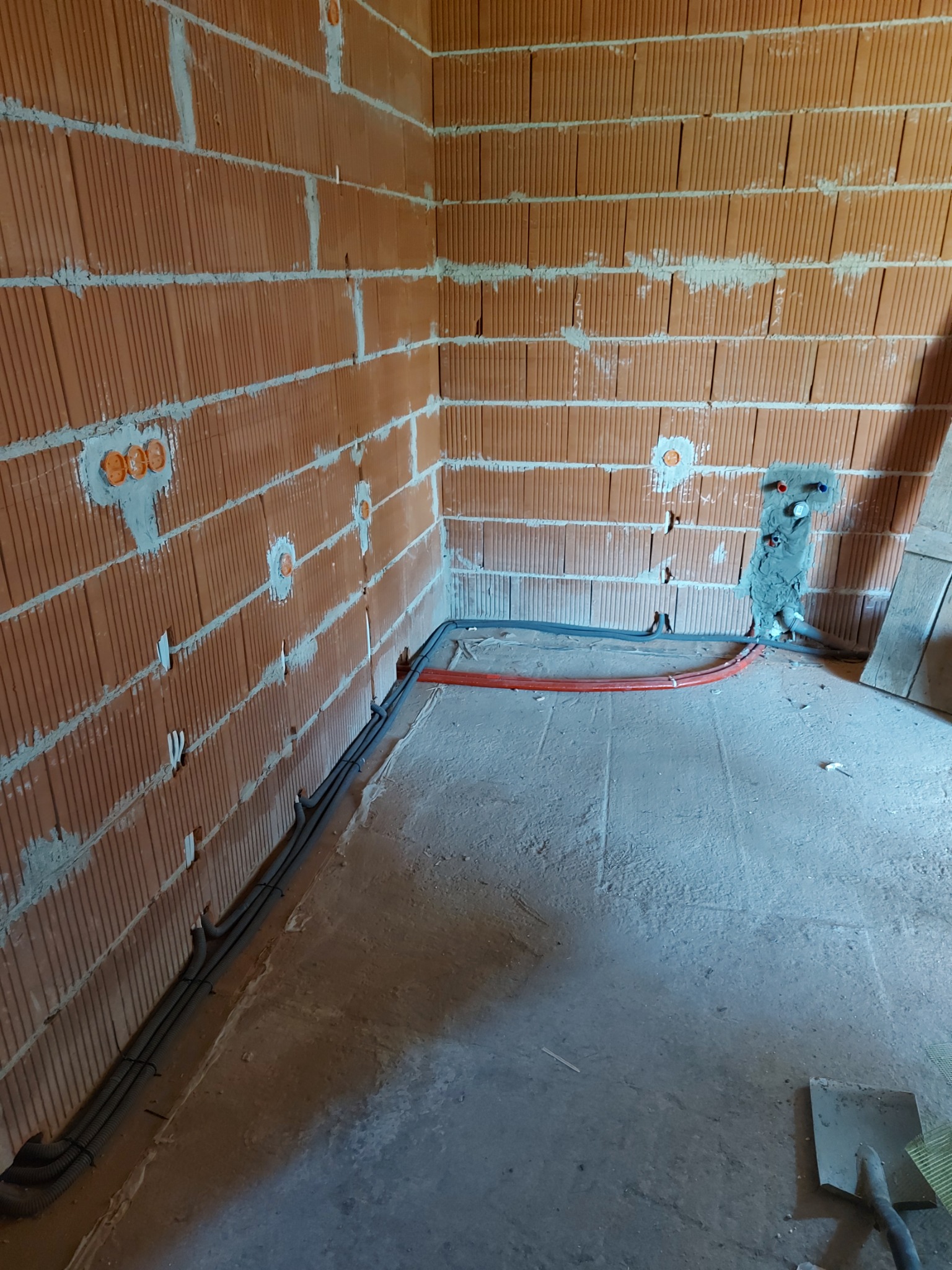 Surowy mur z cegły dziurawki z widocznymi puszkami instalacyjnymi i przewodami elektrycznymi w peszlach, częściowo zamurowane podejście wodno-kanalizacyjne, szary betonowy podkład.