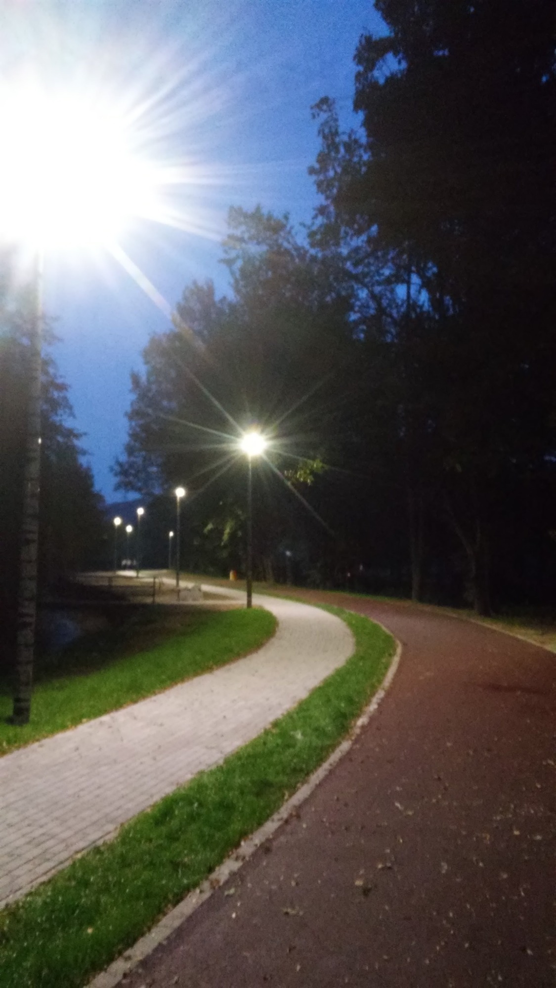 Świecące latarnie parkowe o zmierzchu oświetlają ścieżkę pieszo-rowerową, widoczne drzewa i niebo.