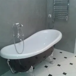 Wolnostojąca wanna na lwich nóżkach z chromowaną armaturą w łazience z szarą ścianą i biało-czarną podłogą w romby, grzejnik drabinkowy na ścianie.