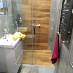 Nowoczesna łazienka z drewnopodobną kabiną prysznicową za szklanymi drzwiami, szara mozaika na ścianach, biała szafka pod umywalką z żółtą ściereczką i czarny grzejnik drabinkowy z różowym ręcznikiem.