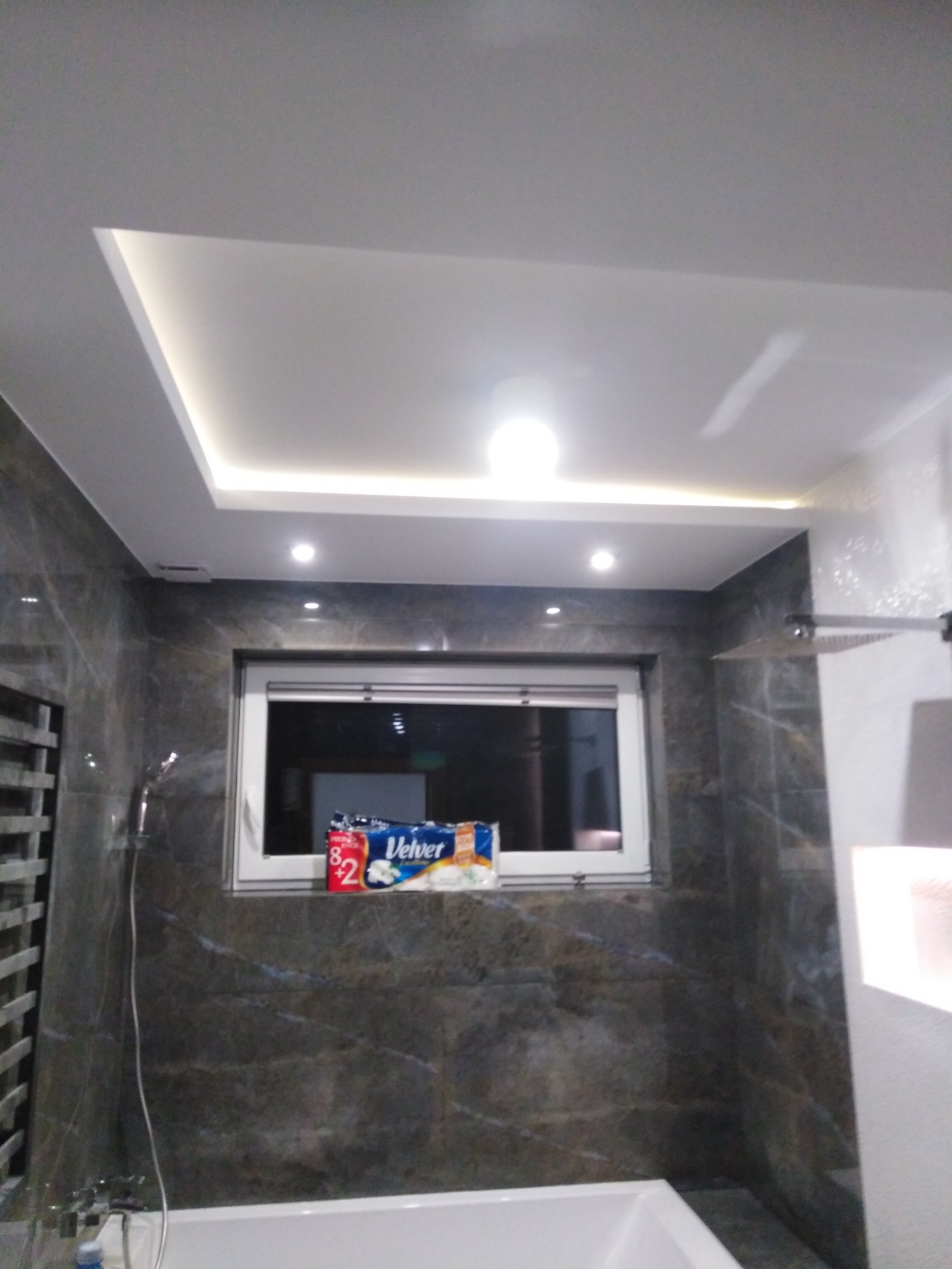 Nowoczesna łazienka z szarymi płytkami imitującymi marmur, podwieszany sufit z oświetleniem LED, biała wanna i okno z widocznymi rolkami papieru toaletowego.