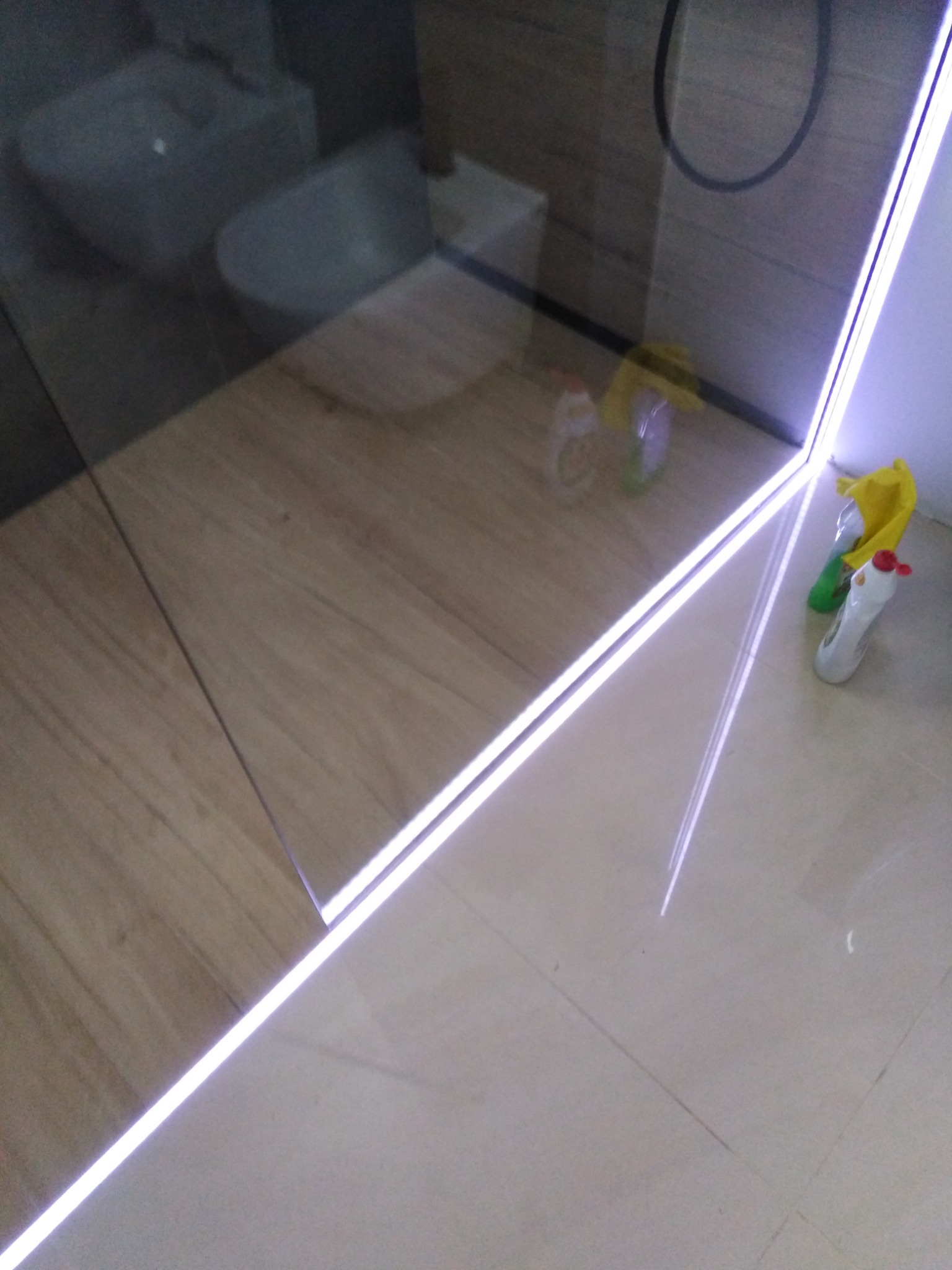 Fragment łazienki z narożną kabiną prysznicową z podświetleniem LED, widoczne płytki imitujące drewno na ścianie oraz jasne płytki podłogowe, na podłodze środki czystości.