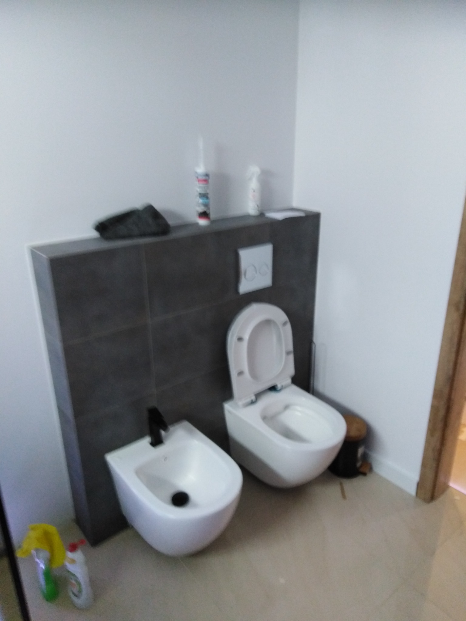 Nowoczesna łazienka z białym bidetem z czarną armaturą i podwieszaną toaletą, wykończona szarą glazurą na ścianie, widoczne środki czystości.