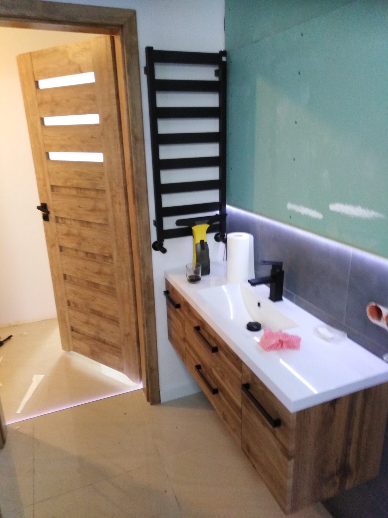 Nowoczesna łazienka w trakcie wykańczania: drewniana szafka z białą umywalką i czarną armaturą, grzejnik drabinkowy, podświetlenie LED, drzwi z przeszkleniami, ściana częściowo wykończona płytami.