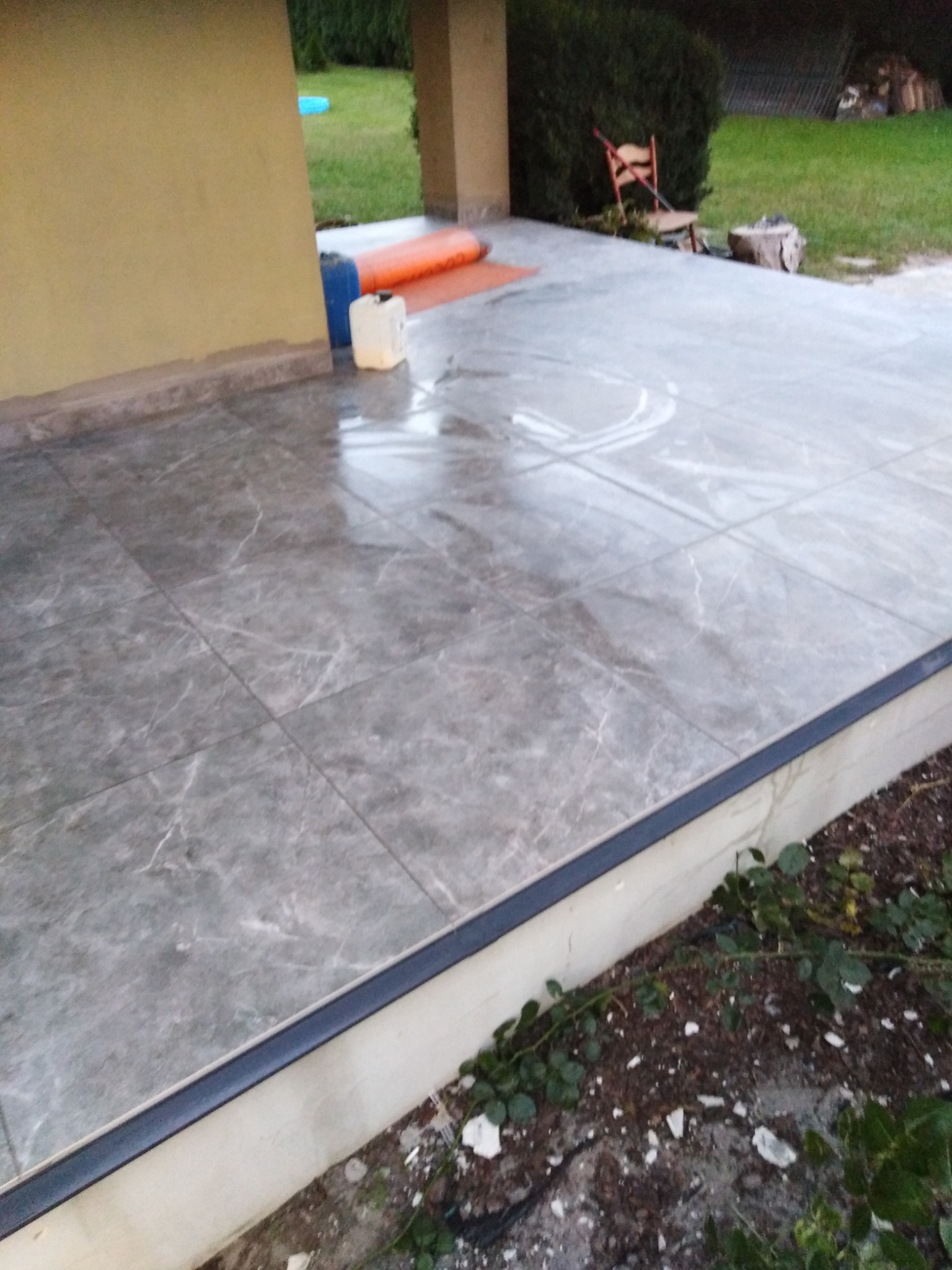 Nowo położona, szara, marmurkowa podłoga z płytek na tarasie, z widocznymi smugami po czyszczeniu, z czarną listwą wykończeniową i fragmentem ogrodu.