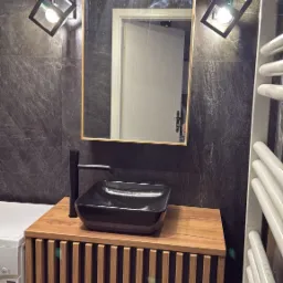 Nowoczesna łazienka z czarną umywalką nablatową, drewnianą szafką z pionowymi listwami, lustrem w złotej ramie i designerskimi lampami ściennymi na tle ściany z ciemnego kamienia.