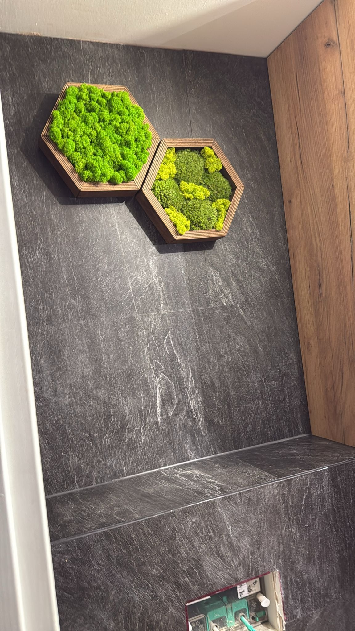 Dekoracja ściany w łazience z dwoma heksagonalnymi ramkami z mchu w różnych odcieniach zieleni, na tle ciemnej, kamiennej płytki i fragmentu drewnianej okładziny.