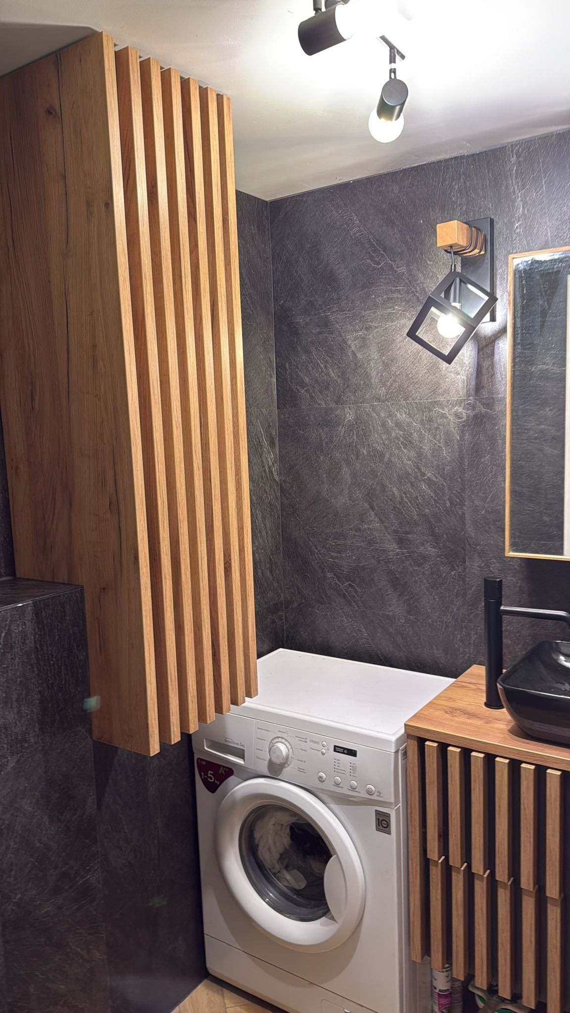 Nowoczesna łazienka z szarą, teksturowaną ścianą, drewnianą zabudową nad pralką i szafką pod umywalkę z lamelami, oświetlona lampą punktową i dekoracyjnym kinkietem.