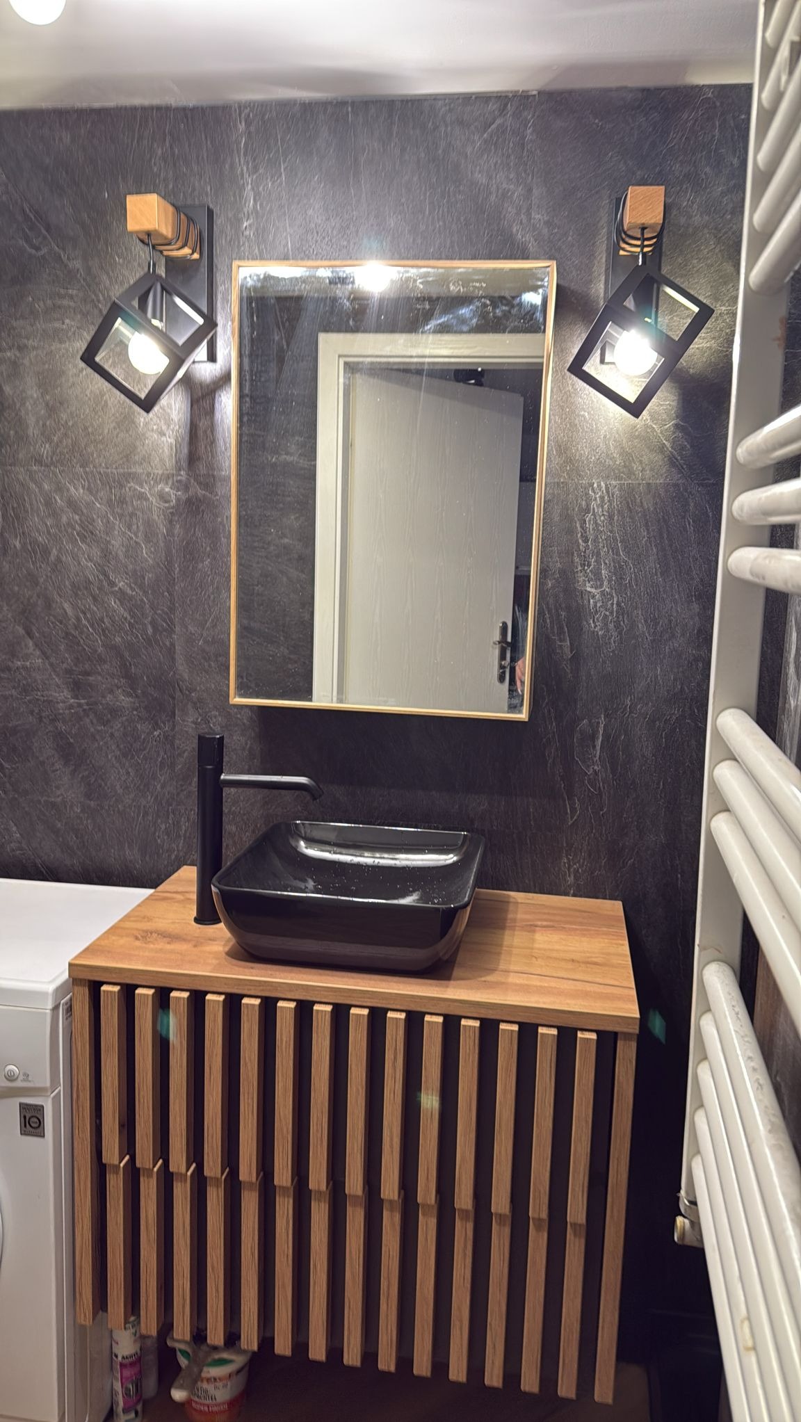 Nowoczesna łazienka z czarną umywalką nablatową, drewnianą szafką z pionowymi listwami, lustrem w złotej ramie i designerskimi lampami ściennymi na tle ściany z ciemnego kamienia.