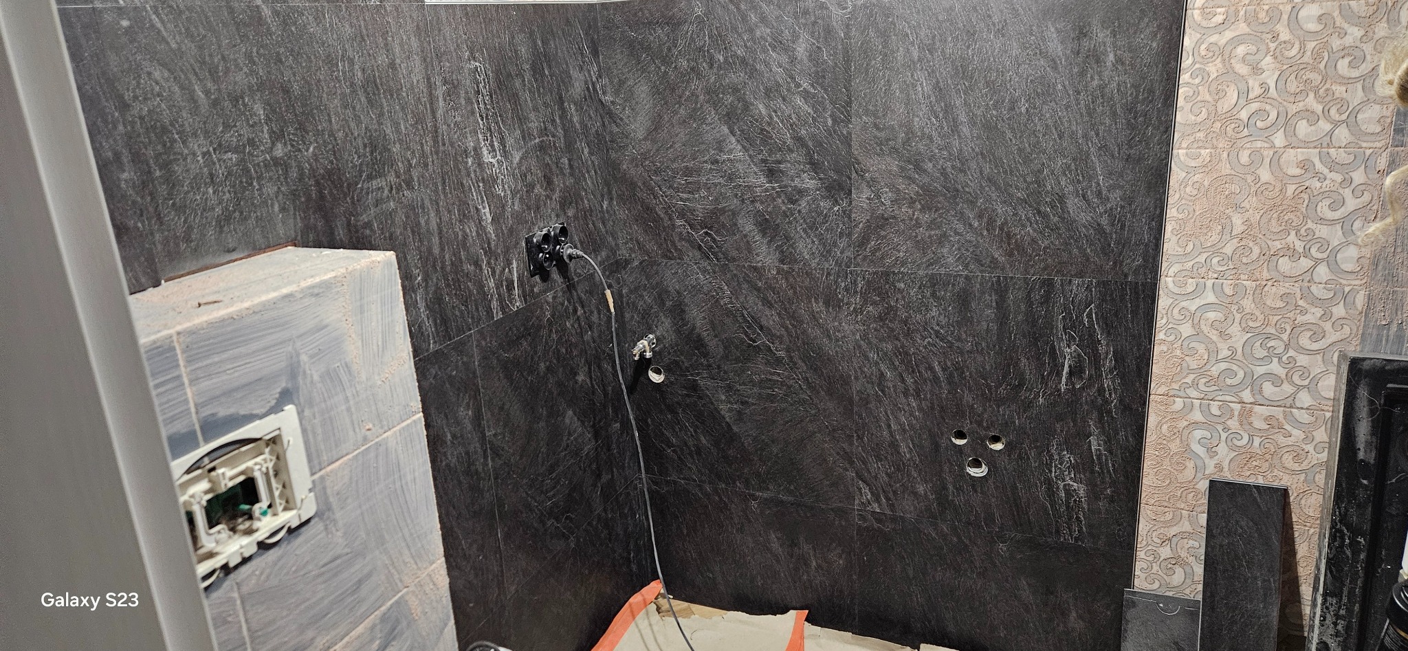 Częściowo wykończona łazienka z ciemnymi płytkami imitującymi kamień na ścianach, widoczne otwory na instalacje, fragment ściany z jasnymi dekoracyjnymi płytkami oraz wystający element instalacji...