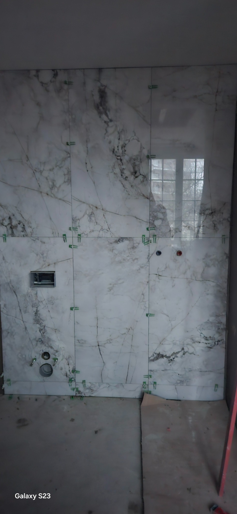 Ściana łazienki w trakcie układania płytek imitujących marmur, widoczne plastikowe krzyżyki dystansowe, otwory na instalacje sanitarne i podtynkowy stelaż WC.