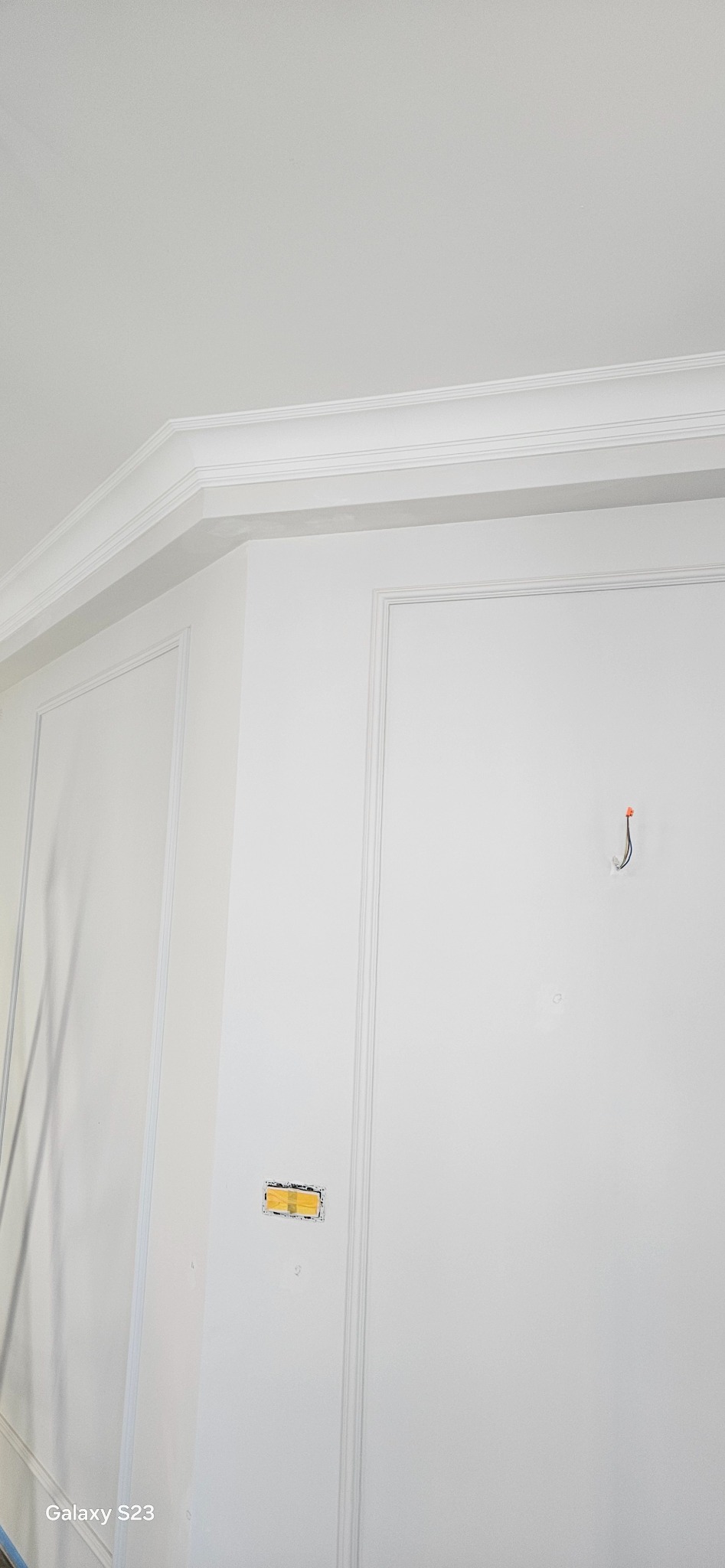 Biała ściana z dekoracyjnymi listwami, gzymsem sufitowym i zabezpieczonym otworem pod gniazdko elektryczne, widoczny haczyk z wystającymi przewodami.
