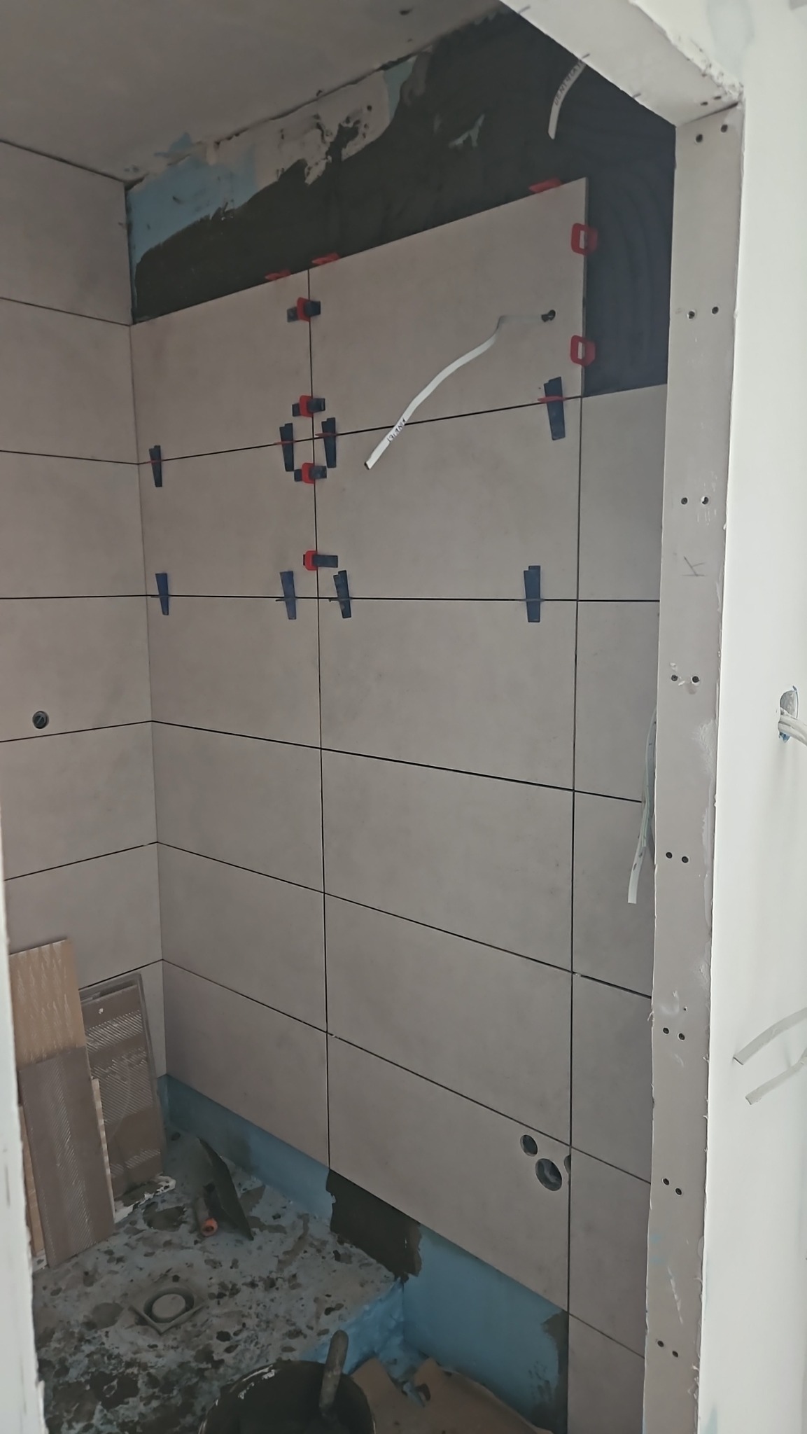 Ściany łazienki w trakcie układania płytek, widoczne poziomice, krzyżyki dystansowe, fragment hydroizolacji i przygotowane otwory na instalacje.