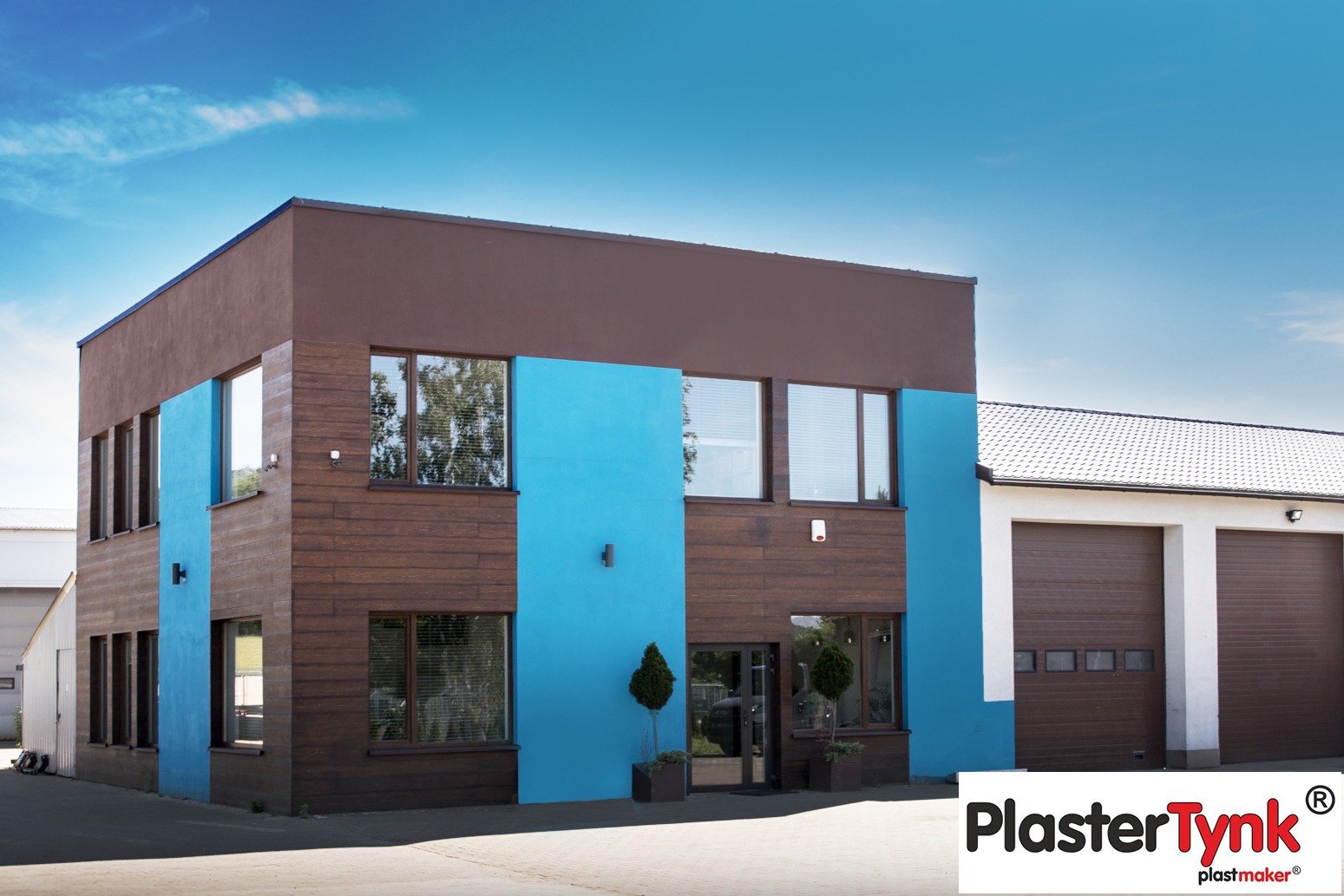 Dwukondygnacyjny budynek z elewacją w kolorach brązu i turkusu, wykończony panelami imitującymi drewno, z widocznym logo producenta tynków.